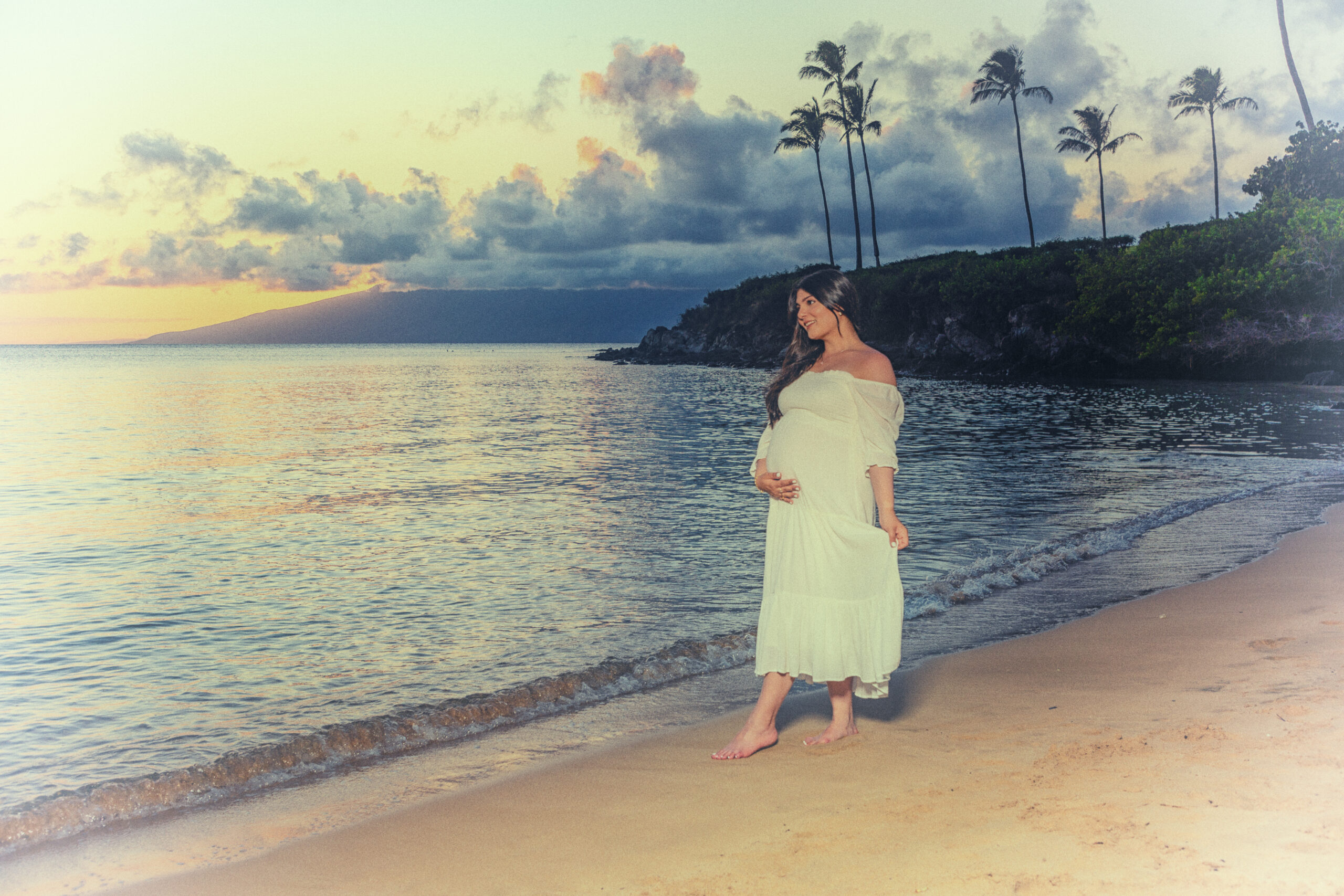 Pregnant woman in white dress by beach at sunset.