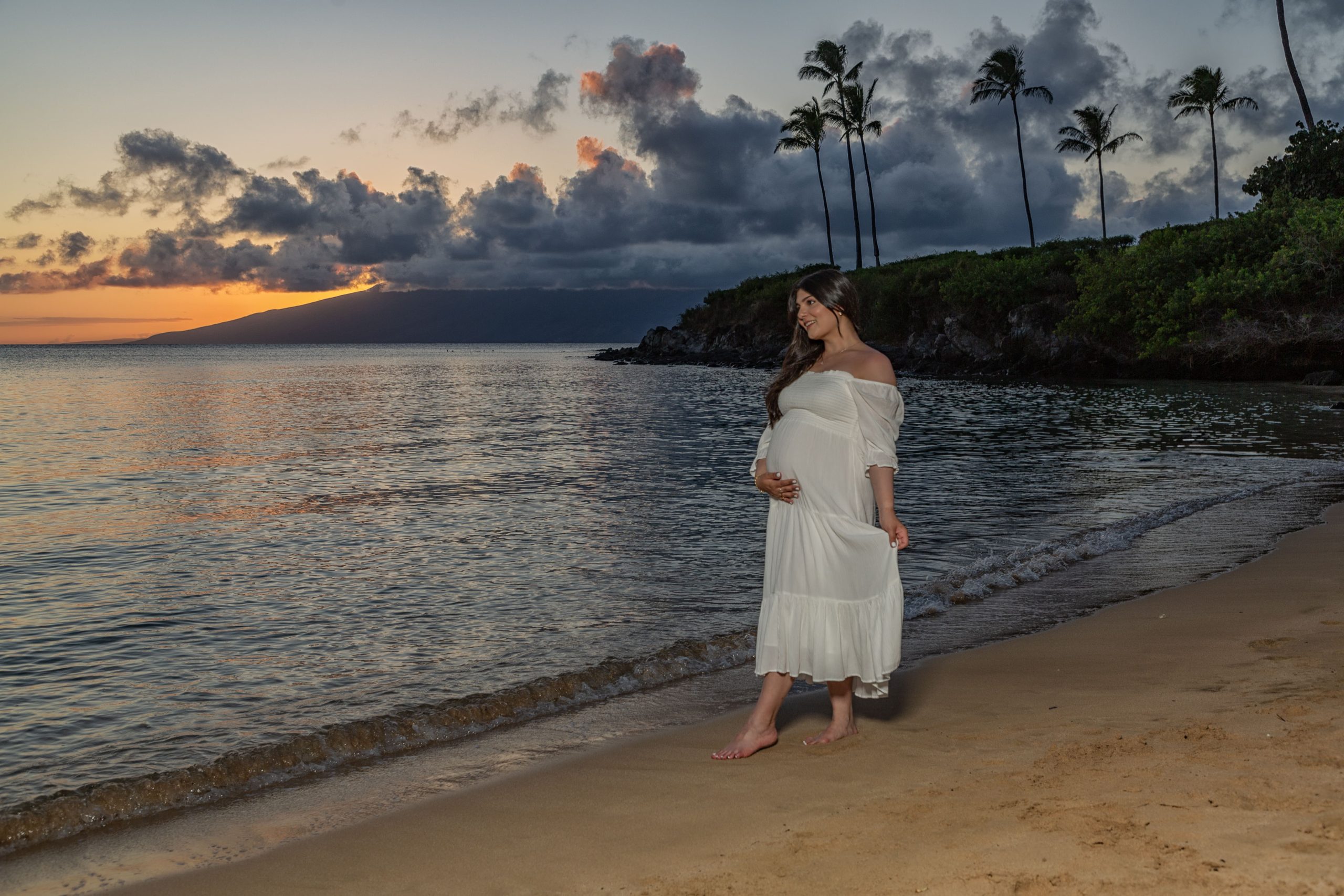 Pregnant woman in white dress on beach at sunset.