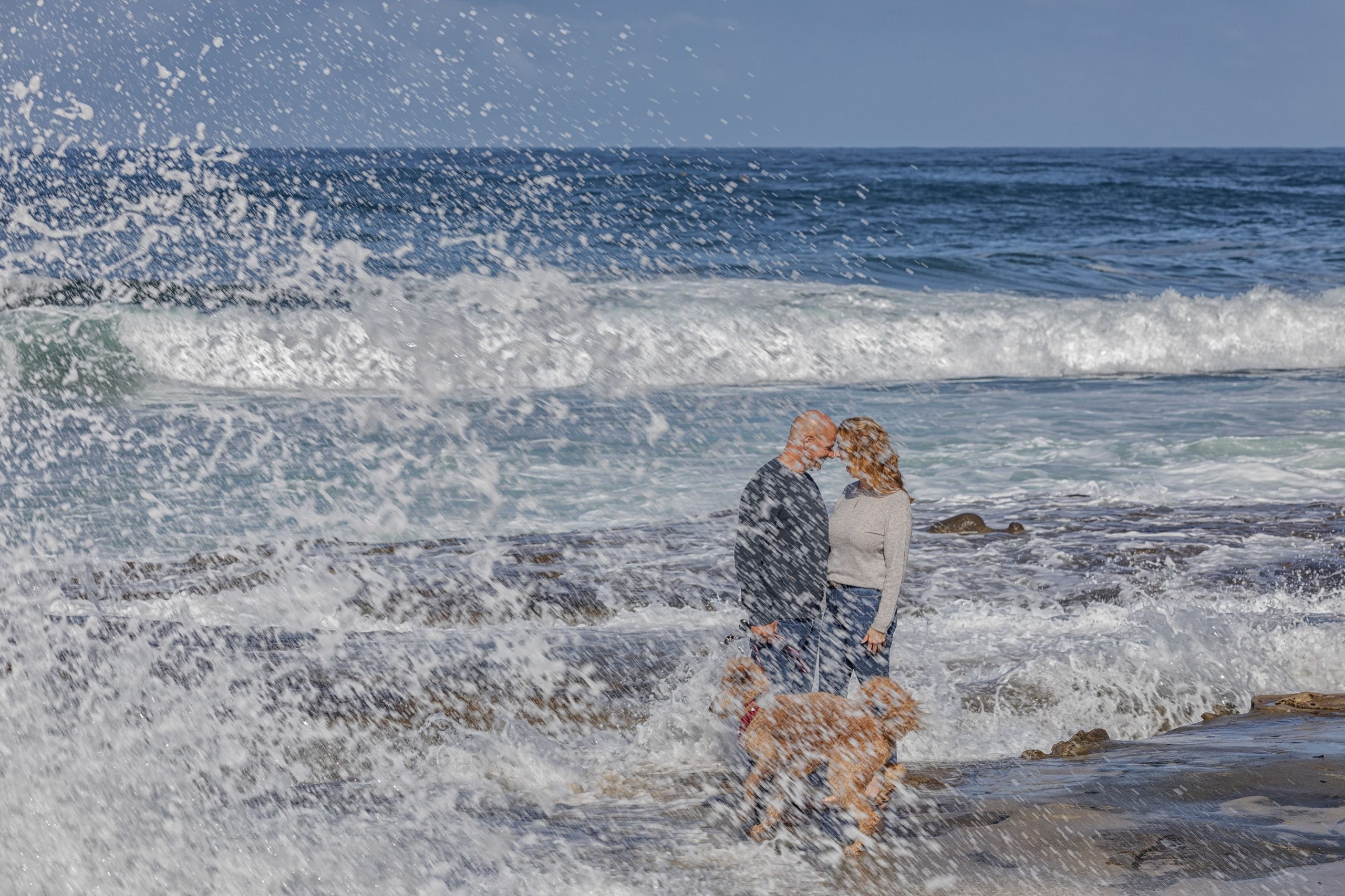 Couple embracing on beach with dog and waves.